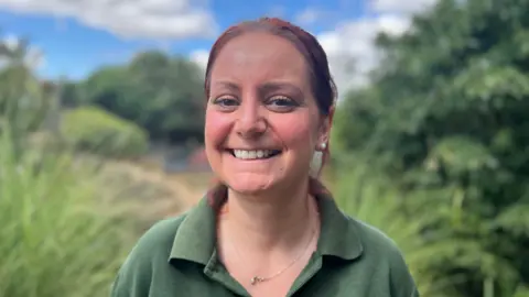 Catherine Doherty wearing a green t-shirt and a necklace and smiling at the camera while in a wooded area