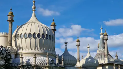 The roof of Brighton's Royal Pavilion. The roof includes three white onion-shaped domes with circular windows as well as pointed tips. The roof is set against a blue sky with clouds.