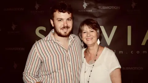 Rachel Prendergast Jonny Hamer wearing a beard and a striped shirt stood in front of a large poster which says Sylvia alongside his mother Heather