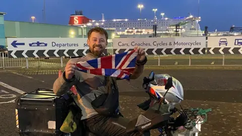 We Hate The Cold Harry Jackson on a moped, by a large passenger ferry, at Harwich docks. He is holding up a union jack and has a helmet on his handlebars. He is smiling and has short dark hair and a beard. 