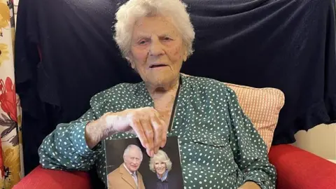 Ethel Caterham sat holding a card with a picture of the King and Queen on the cover.