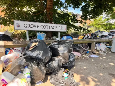 A number of bins bags are on the pavement along with other rubbish by the road sign.
