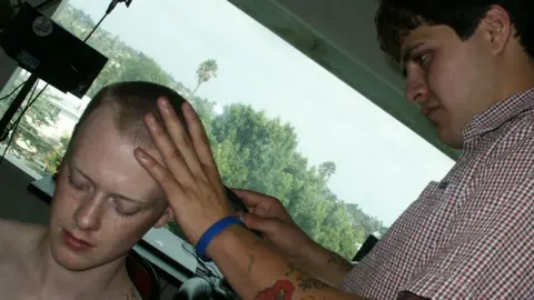 Olivia Osby Un hombre sentado mientras le cortan el pelo. Está en la esquina inferior izquierda de la imagen, que está inclinada. Su cabello ha sido afeitado al estilo rapado. Otro hombre que lleva una camisa estampada se está cortando el pelo. Ese hombre tiene cabello castaño y sostiene la navaja en una mano con la otra colocada en la cabeza del primer hombre. Se puede ver una ventana al fondo que revela un día brillante en Los Ángeles lleno de árboles.