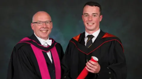 Father and son John and Cameron Gibson, pictured inside at Cameron's graduation. Both are wearing gowns and Cameron is holding his degree.