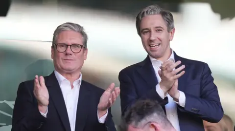 Niall Carson/PA Wire Sir Keir Starmer and Simon Harris, wearing dark suits and open neck shirts, standing and applauding in Dublin's Aviva stadium 