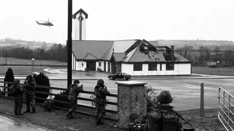 Pacemaker Five soldiers wearing camouflage gear stand guard beside the carpark of St Patrick's Church, which has a partially crumbling rook. A car sits stationary in the car park while a helicopter flies overhead. 