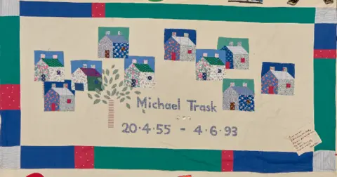 UK Aids Memorial Quilt picture of a quilt that has houses and a tree stitched on as well as the name Michael Trask