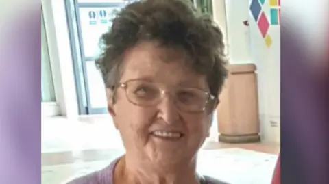 Celia Adams is pictured smiling at the camera wearing glasses and a purple top. She has short brown hair.