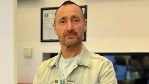 BBC Mark Bishop stood in the BBC building. He is smiling at the camera and wearing a light denim jacket. 
