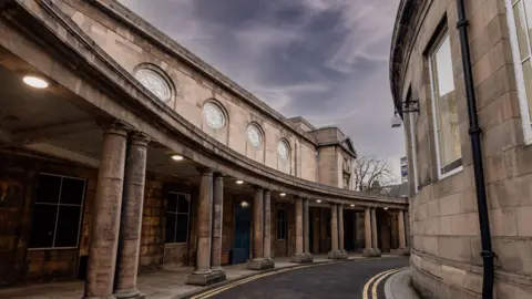 A curved road outside a sandstone building with a columned walkway and round windows above it