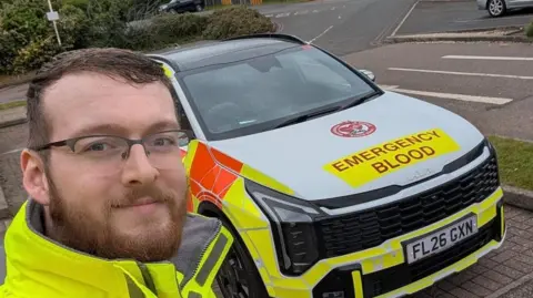 A man dressed in hi vis jacket stood infront of an emergency vehicle, with yellow signage 