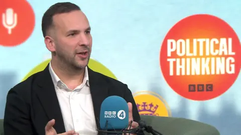 Green Party of England and Wales leader Zack Polanski speaking in the Political Thinking studio. He is wearing a black suit jacket and a white shirt with an open neck. 