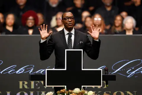 Getty Images Former basketball player Isaiah Thomas has two hands up as he speaks from the podium at the service. He is wearing a suit and a pol-a-dot tie.