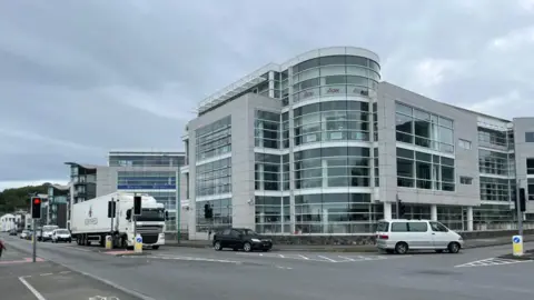 Office buildings housing finance businesses next to a road with cars, vans and lorries on the road.