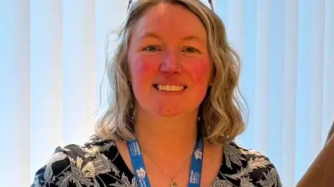 Marion McLaughlin standing in front of closed blinds. She is wearing a black and grey dress and a blue lanyard. She is wearing her glasses on her head and has short blonde hair.