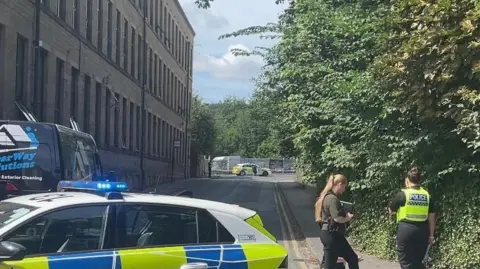 Yappapp A three storey building on the left next to a road. A police car in the foreground and another at the end of the road. Two female officers stand on the road.