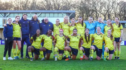 Lucy Copsey A football team photo. The players and staff are standing or crouching down on a football pitch. The players are wearing a yellow kit and the staff are wearing a blue kit.