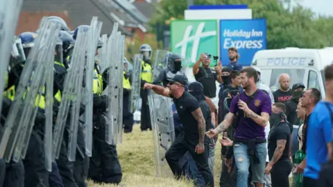 A picture of the disorder outside the Holiday Inn hotel in Rotherham. It shows a line of police officers wearing helmets and holding shields who are confronted by a group of men.