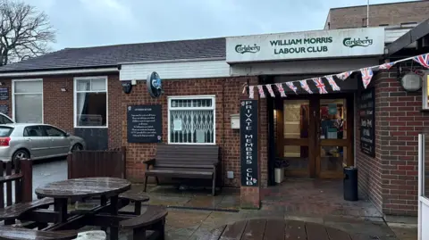 Martin Heath/BBC Exterior shot of the club, with a white board above the door showing WILLIAM MORRIS LABOUR CLUB in green lettering. There is union jack bunting over the lintel and a vertical sign to the left of the door states PRIVATE MEMBERS CLUB. Two wooden tables with benches around them are visible in the foreground and a car is parked to the left.