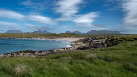 Getty Images Clear blue waters of the sea and a beach with sand and rocks. In the distance are mountains under a blue sky with clouds.