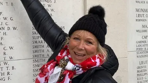 Sadie Nine, wearing a black hat, with a red patterned scarf, black coat, with short fair hair, pointing at a war memorial - a large grey monolith with dozens of names engraved on it.