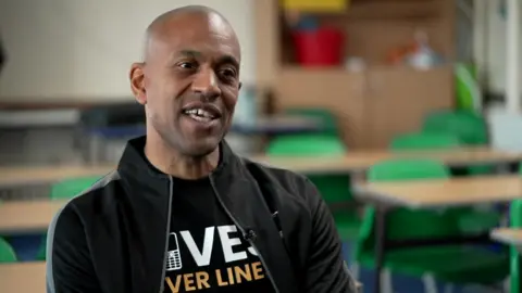 A bald man sits in an empty classroom in front of desks and green plastic chairs