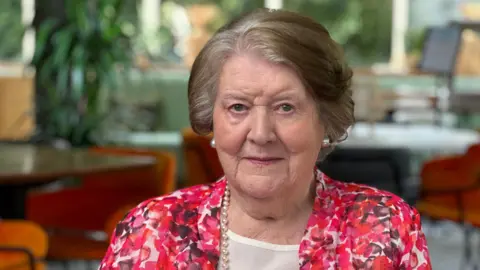 Patricia Routledge sits indoors wearing a pink, red and white patterned jacket and a pearl necklace. The photograph was taken in a bright room with large windows and orange chairs visible in the background.