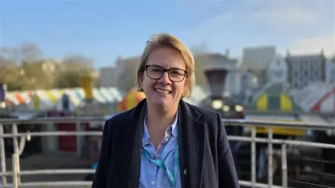 Paul Moseley/BBC Carli Harper stands in front of Norwich market. She is in a dark coat over a blue shirt. She has cropped blonde hair, is wearing dark glasses and smiling at the camera.