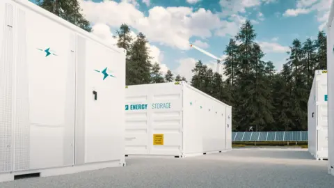 Large white battery storage units in a forest landscape with solar panels and a wind turbine in the background