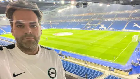 Barry Hill A selfie of Barry Hill at the Amex Stadium in Brighton. He is wearing a white Brighton & Hove Albion football shirt. The green pitch and blue stands are in the background. 