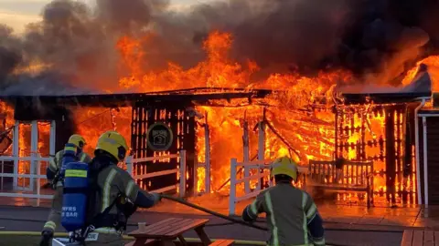 A close up picture of the flames ripping through the cafe at the Old Sarum Airfield. It shows a one-story building engulfed in bright flames and three firefighters in the foreground stretching a hose towards the fire.