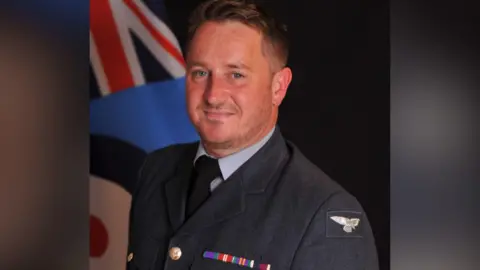 A man is smiling proudly into the camera. He has short light brown hair and is wearing a blue suit, with dark blue tie. The suit has RAF wings on the left shoulder and behind him is a flag of Great Britain.