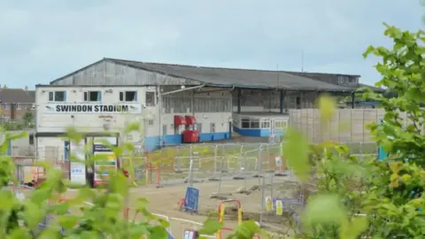 LDRS The edge of Swindon Abbey Stadium. There is a white and blue building with a metallic roof. At the side of the building hangs a white banner with black writing reading 'Swindon Stadium'. There are silver fences joined together in front of the stadium.