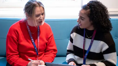 Northamptonshire Police, Fire and Crime Commissioner One woman with her light coloured hair tied up, wearing a red jumper and a pink, purple and blue lanyard, holding a pen and notepad speaking to another woman. The second woman is also wearing the same lanyard, with her dark curly hair also tied up and she's typing on a laptop. 
