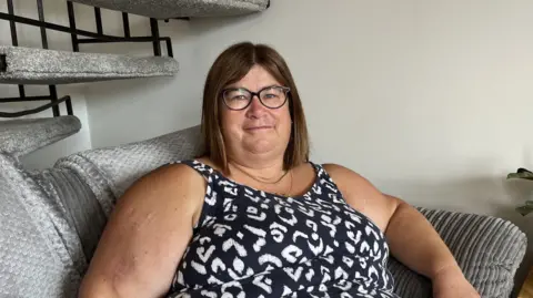 Shaun Whitmore/BBC Allison Heathcote is wearing a black and white patterned dress and black glasses. She is sitting on a grey sofa and looking directly at the camera.