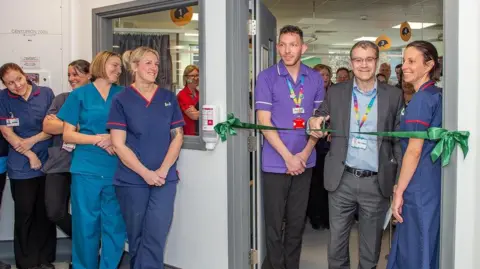 Craig Hobbs photography A group of people wearing scrubs smiling as a man in a suit cuts the ribbon at the entrance to a hospital ward.