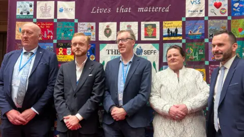 Five people standing in a row. Four men are wearing blue suits and a woman is wearing a cream jacket.