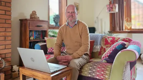 A man sat on a sofa in front of a computer, wearing an orange jumper, blue shirt and beige trousers. There are colouful throws and cushions behind him, along with wooden furniture and wooden framed windows. 
