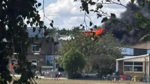 Clare Perkins A distant shot shows flames above tree tops and black smoke billowing into the sky