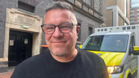 A man with grey hair and grey facial stubble with black glasses, wearing a black top. Behind him is an ambulance vehicle that is yellow and white.