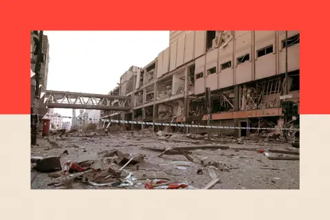PA Media A photo dated 16 June 1996 shows the scene of devastation in Manchester City Centre following the bomb attack. Police tape stretches across the street and rubble is strewn over the road. Behind is the Arndale shopping centre. Its windows have been blown out.