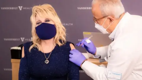 Reuters Dolly Parton wears a blue top and a matching face mask as a doctor in a white coat and purple latex gloves prepares to inject her shoulder. 