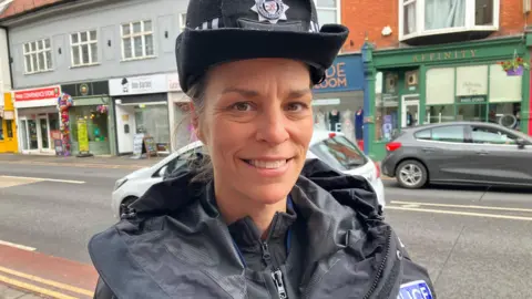 A female police officer in uniform standing by the side of a high street smiling at the camera