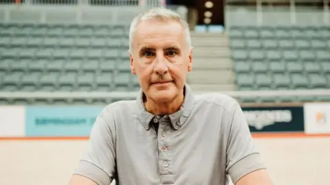 Getty Images Dermot Murnaghan looks into the camera while wearing a grey t-shirt in an arena in Birmingham in 2022.