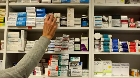 PA Media A woman's arm is reaching across shelves packed with boxes and bottles of medicines.