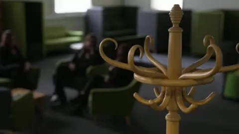 A blurred image of three women sitting around a coffee table, a hat stand is in focus in the foreground and other sofas are visible in the background.