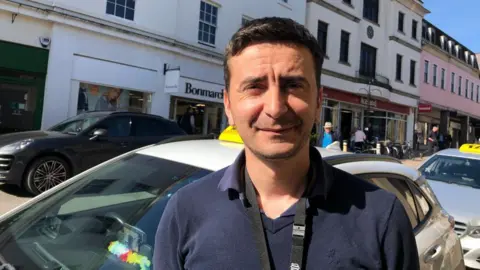 Vikki Irwin/BBC Alin Mortoiu is standing outside his Taxi and on the street in Bury St Edmunds. He is wearing a blue polo shirt, has dark hair and is smiling at the camera. 