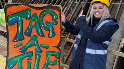 Cornerstone A woman, wearing a hard-hat, with a blue coat and a blue high-vis jacket, pointing upwards, holding a large sign, in orange and green, that says Tag-A-Tile. A roof if behind her, with slates missing from it. 