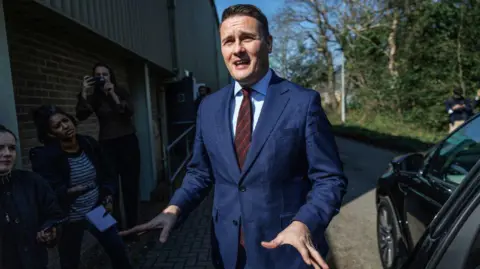 Wes Streeting stood in front of a crown, wearing a suit. 