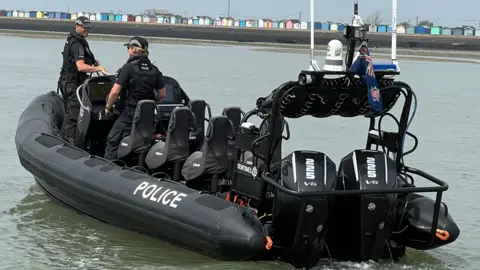 A wide shot of a black boat out on the water with the word police written along the side. Two officers are on board